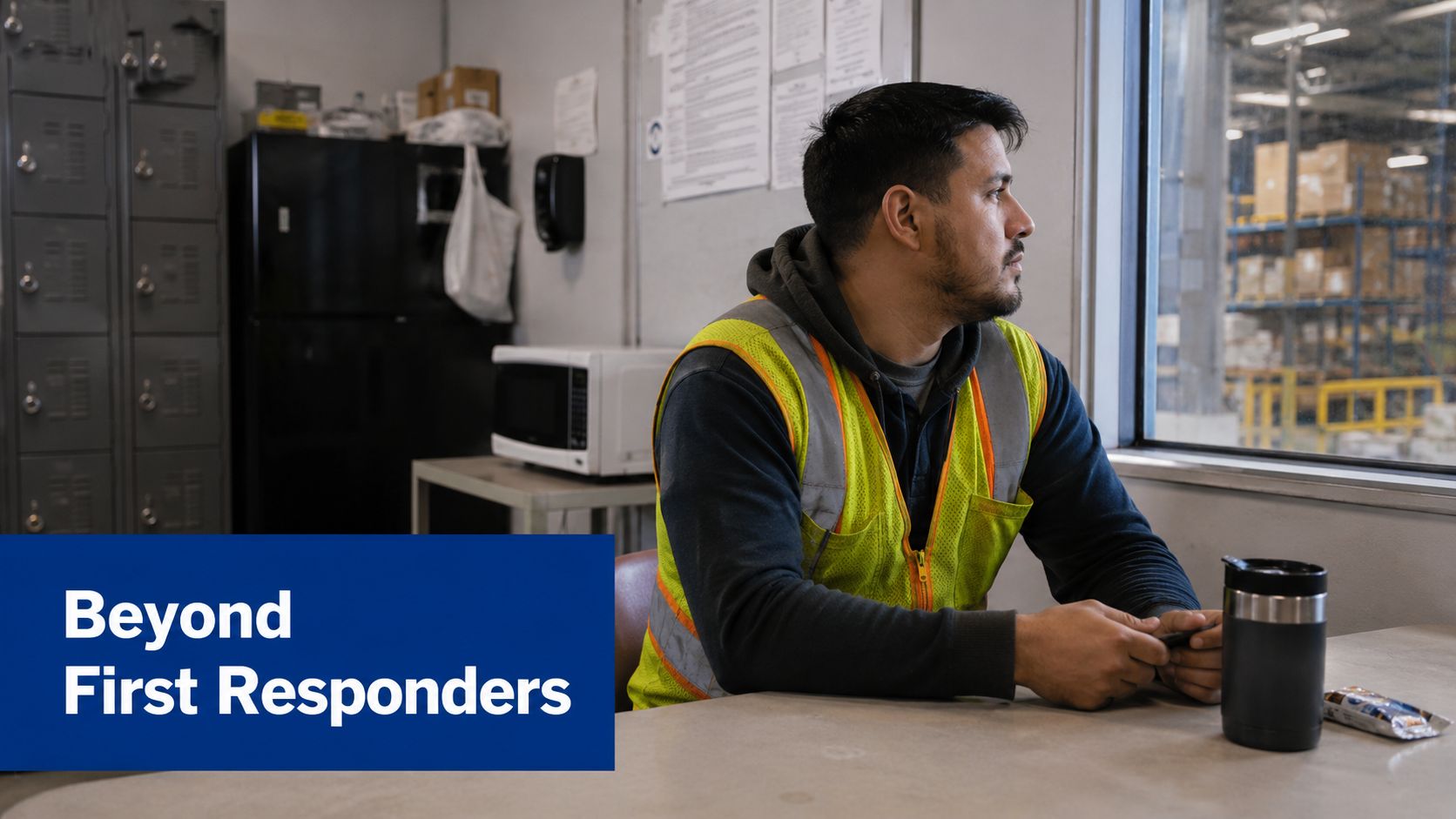 A warehouse worker sitting in a breakroom, looking out the window while holding a cell phone.